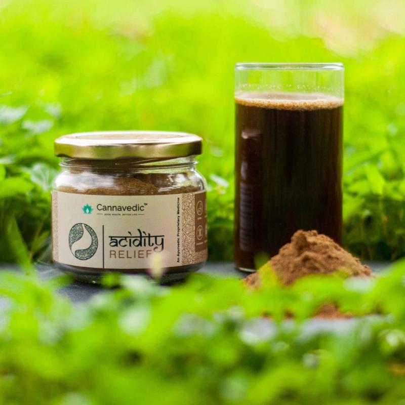 Jar of "Cannavedic Acidity Relief" powder next to a glass of dark liquid and a small mound of the powder in a green, leafy background.