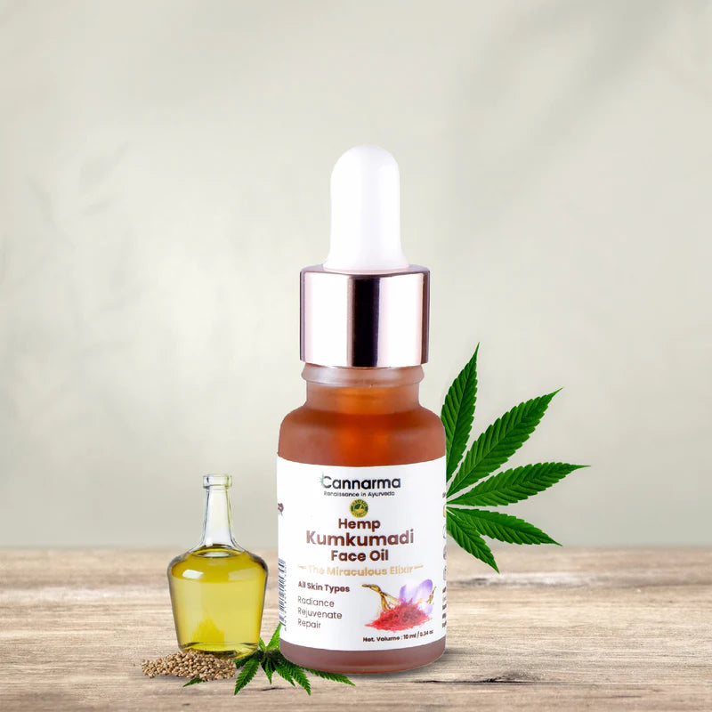 A brown glass bottle of Cannarma Hemp Kumkumadi Face Oil with a white dropper cap, besides hemp seeds, and hemp leaves.