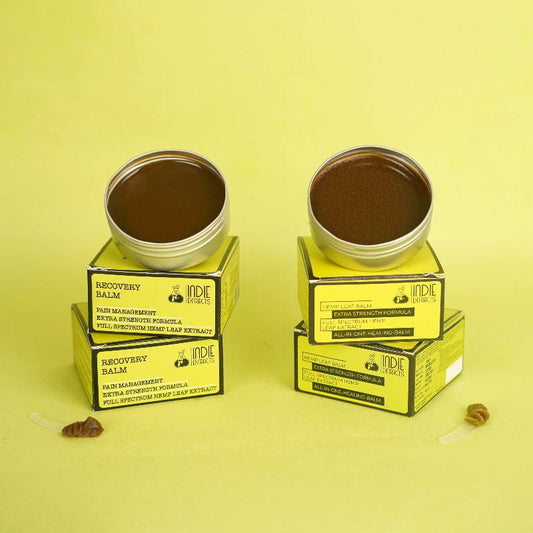 Two open tins of balm on yellow boxes, labeled "Recovery Balm" and "Hemp Leaf Balm," on a light yellow background.