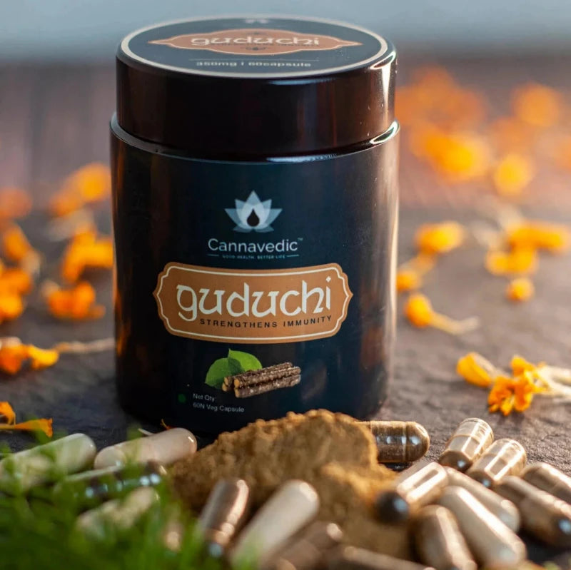 Alt-Text: Black container of Cannavedic Guduchi capsules, surrounded by capsules, powder, and flower petals.