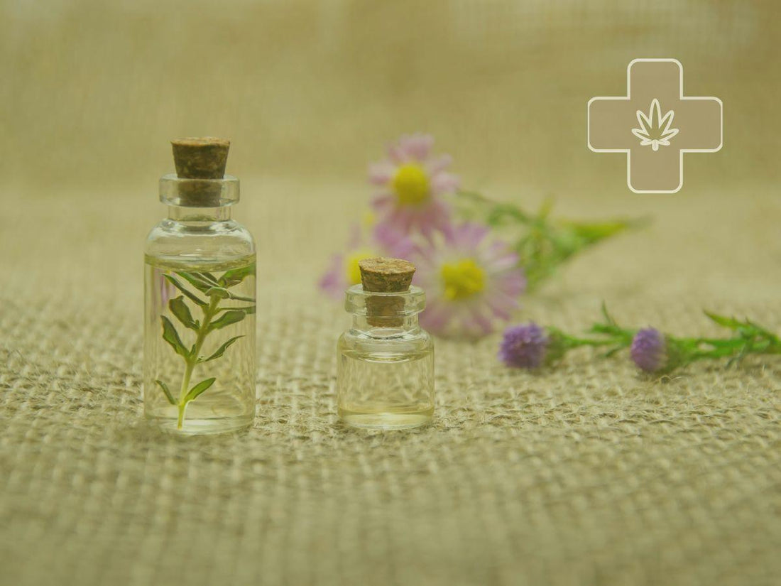 Two glass bottles with corks on burlap, one with green leaves inside, with blurred purple and pink flowers in the background and a medical cross symbol with a leaf.