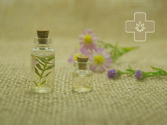 Two glass bottles with corks on burlap, one with green leaves inside, with blurred purple and pink flowers in the background and a medical cross symbol with a leaf.
