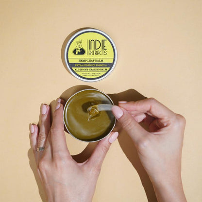 Indie Extracts hemp leaf balm tin open on beige surface, with hand scooping greenish balm using a small tool.