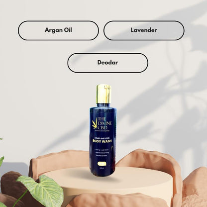 A dark blue bottle of "THE DIVINE CBD" body wash on a beige pedestal, with text boxes above reading "Argan Oil," "Lavender," and "Deodar."

