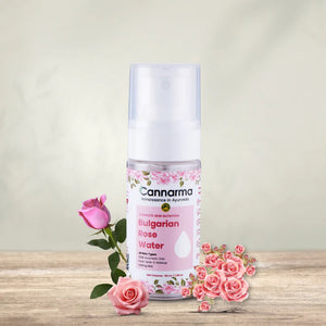 Cannarma Rose Water Oil spray bottle on wood, surrounded by pink roses. For all skin types, with vitamin C.