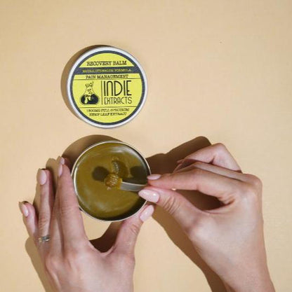 Hands scooping Indie Extracts CBD balm from a tin with a yellow label on a beige background.
