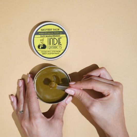 Hands scooping Indie Extracts CBD balm from a tin with a yellow label on a beige background.