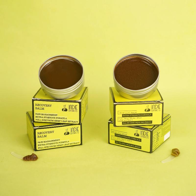  Two open tins of balm on yellow boxes, labeled "Recovery Balm" and "Hemp Leaf Balm," on a light yellow background.