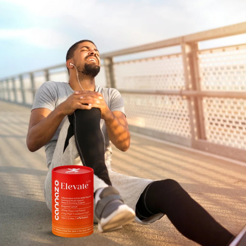 A man sitting on a bridge railing at sunset stretches his leg, wearing a gray t-shirt and black leggings. A red cylindrical canister of Cannazo Elevate CBD oil is placed in the foreground on the pavement.