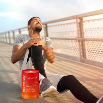 A man sitting on a bridge railing at sunset stretches his leg, wearing a gray t-shirt and black leggings. A red cylindrical canister of Cannazo Elevate CBD oil is placed in the foreground on the pavement.