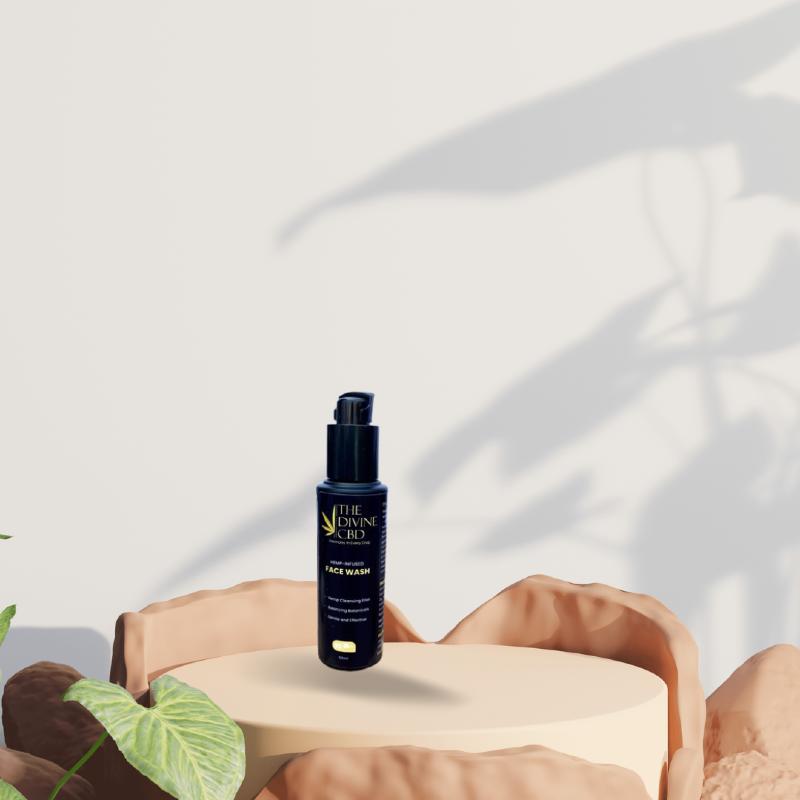 Black bottle with pump dispenser labeled "The Divine CBD Face Wash" on beige pedestal with leafy shadows in the background.

