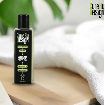 Bottle of Cure By Design Hemp Seed Oil on a wooden surface with a white towel and green leaves in the background.