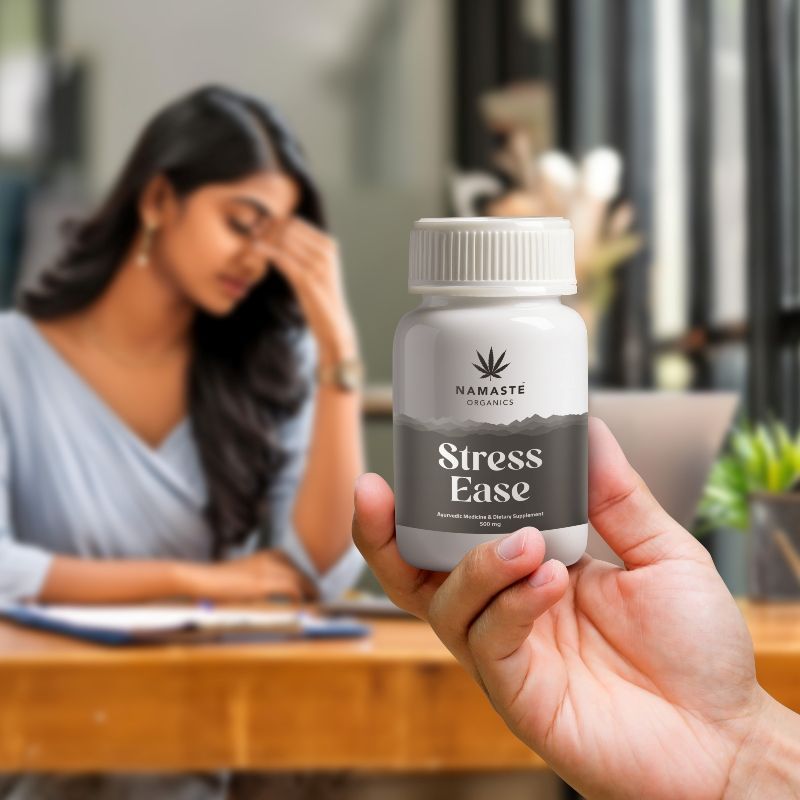 Hand holding a bottle labeled 'Stress Ease'  Full spectrum CBD Capsule by Namaste Organics with a blurred background of a woman sitting at a desk.