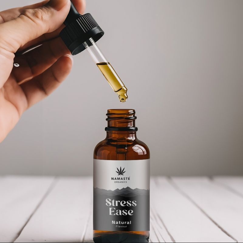 Hand holding a dropper over a bottle labeled 'Stress Ease' Full spectrum CBD Oil by Namaste Naturals on a wooden surface.