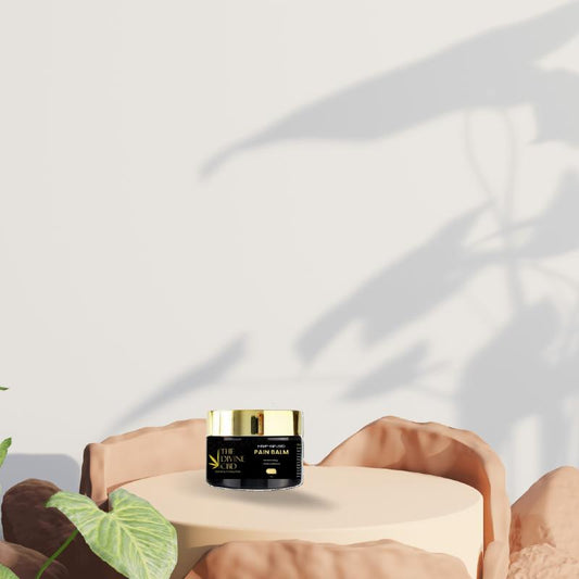 A jar of "THE DIVINE CBD PAIN BALM" on a cylindrical platform, with green leaves and rocky elements in the background.

