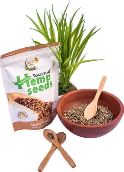 Packaged toasted hemp seeds with a bowl of seeds, wooden spoons, and a plant on a white background