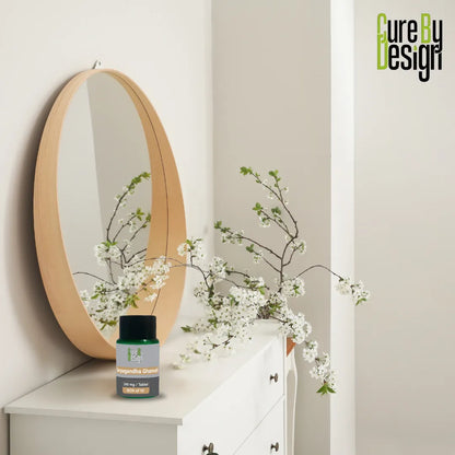 Round mirror on a shelf with white flowers and a bottle labeled 'Cure by Design' against a light gray wall.