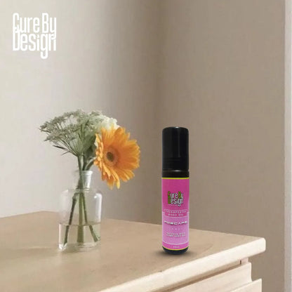 Therapeutic Body Oil PMScape by Cure by Design bottle with purple label on a surface next to a vase with flowers, against a plain wall.