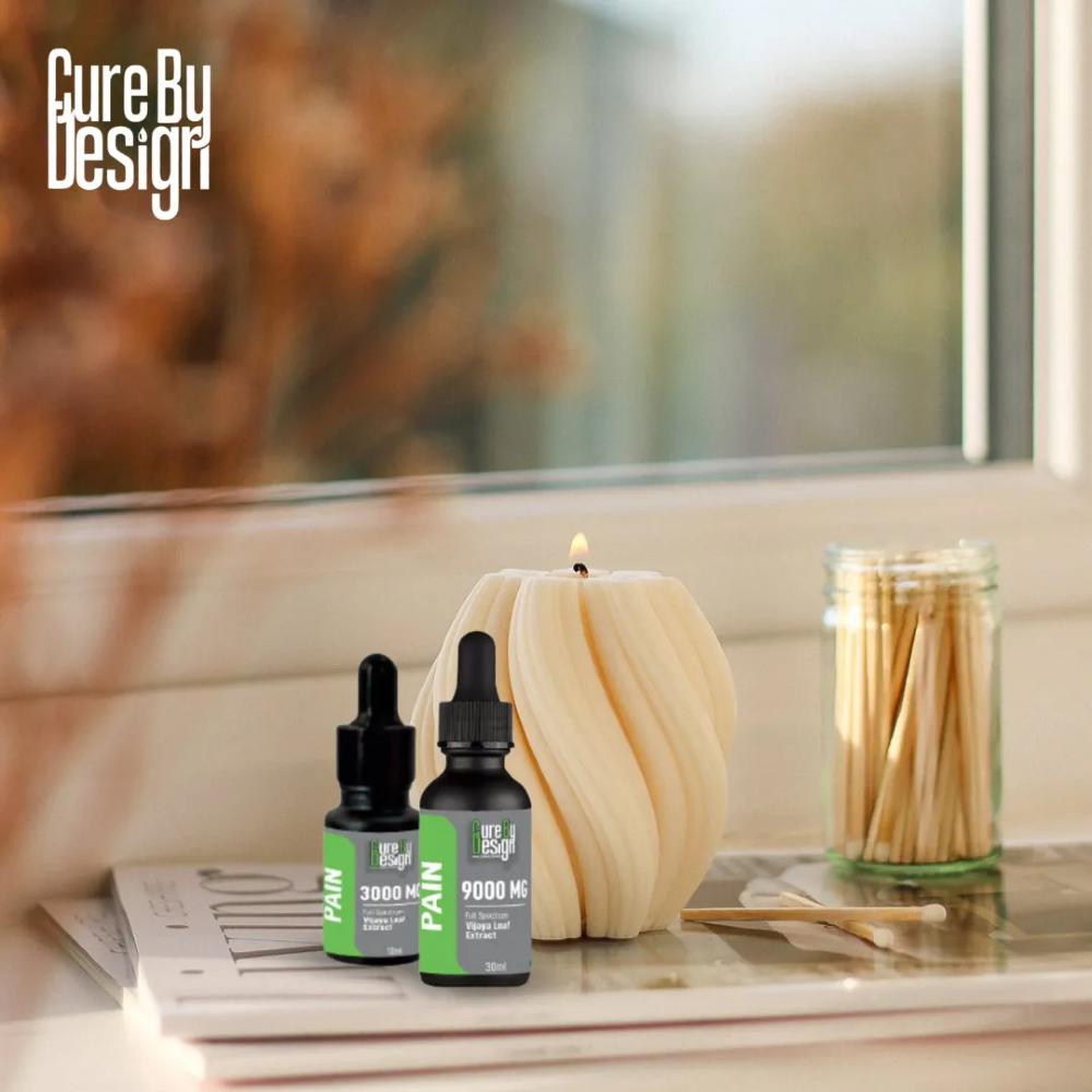 Two bottles of 'Cure By Design' pain relief product on a surface with a candle and matches in the background.