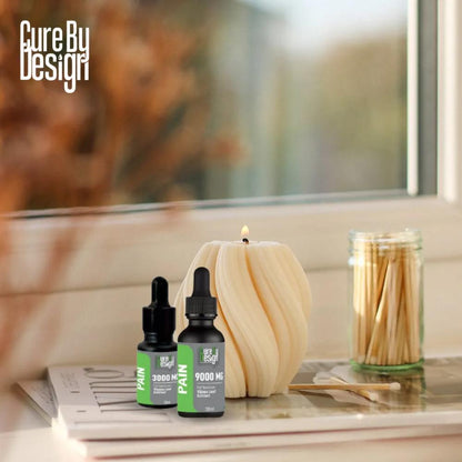 Two bottles of 'Cure By Design' pain relief product on a surface with a candle and matches in the background.