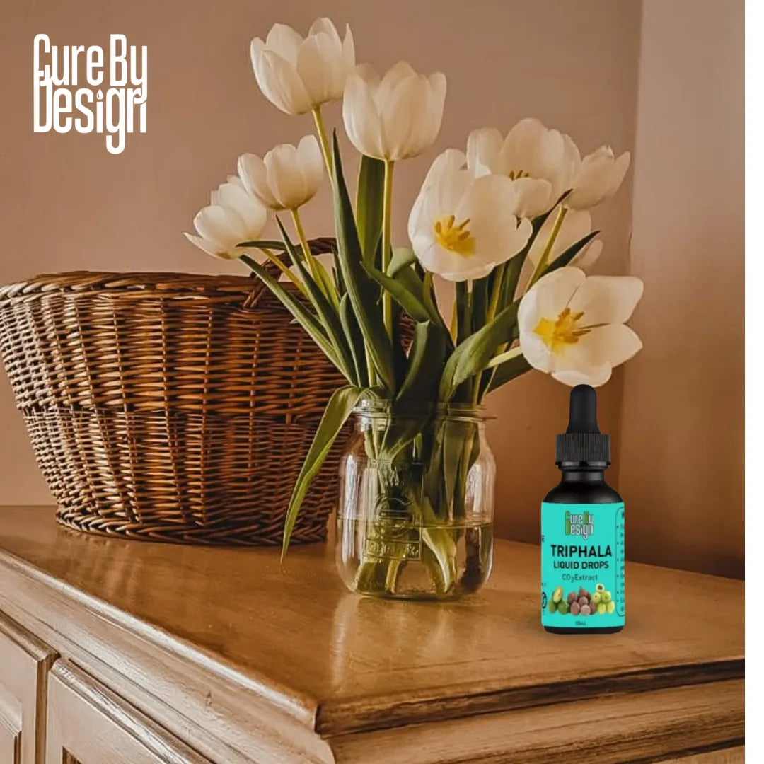 Bottle of Cure By Design Triphala liquid drops on a wooden surface with a vase of white tulips and a wicker basket in the background.
