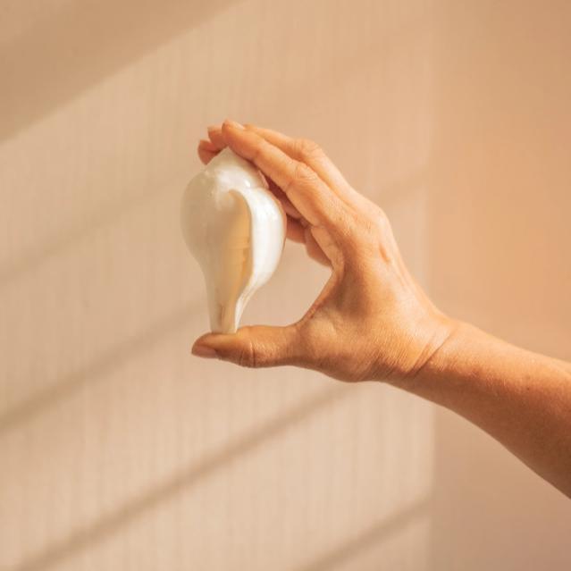 A hand holding a smooth, white conical shell (Shankh) against a sunlit beige background.